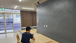 A person sitting at a wooden table in a room with a grey textured wall and a window with curtains. - Guest house cocode. - Hostel (Ishigaki)