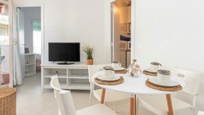 Una habitación moderna y bien iluminada, con un televisor sobre un mueble blanco, una pequeña planta en maceta, y una mesa de comedor con sillas blancas y individuales tejidas.