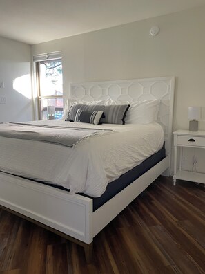 Una cama bien tendida con ropa de cama blanca y un colchón azul oscuro, una mesita de noche blanca con una lámpara, y un piso de madera.