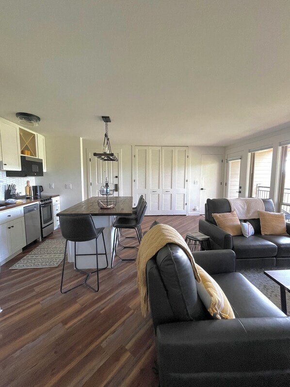 Una cocina moderna con armarios blancos, un comedor con mesa y sillas negras, y una sala de estar con un sofá negro y una mesa de centro.