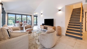Una sala de estar moderna con un televisor de pantalla plana, asientos cómodos, una escalera y ventanas amplias que ofrecen vistas al exterior.