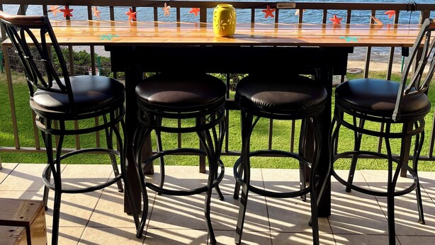 Een houten tafel met vier zwarte stoelen, een gele vaas en uitzicht op zee en bergen.