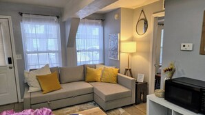 A living room with a sofa, a table with a plate and flowers, a lamp, a TV, and a microwave. - The Port - Walkable to University of Florida, includes Parking (Gainesville)