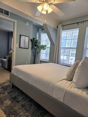 A bedroom with a large bed, a ceiling fan, a window with blinds, and a plant. - The Port - Walkable to University of Florida, includes Parking (Gainesville)