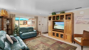 A living room with a large flat-screen TV, a blue leather sofa, a wooden entertainment unit, and a view of the outdoors through a glass door. - AC Family friendly 3 bed/3 bath, Spacious, Comfortable, AC, WiFI, Beach Toys and Parking (Newport Beach)