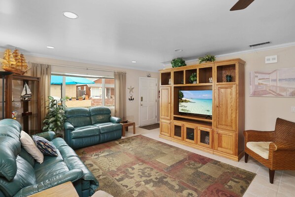 A living room with a large flat-screen TV, a blue leather sofa, a wooden entertainment unit, and a view of the outdoors through a glass door.