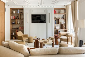 A living room with a sofa set, two armchairs, a coffee table, and a TV. There are bookshelves on both sides of the room. - Merveil - Signature Suite - Marais - Caire II (Paris)