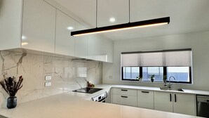 Une cuisine moderne avec des meubles blancs, un pare-feu en marbre, une cuisinière encastrée et une fenêtre avec des stores.