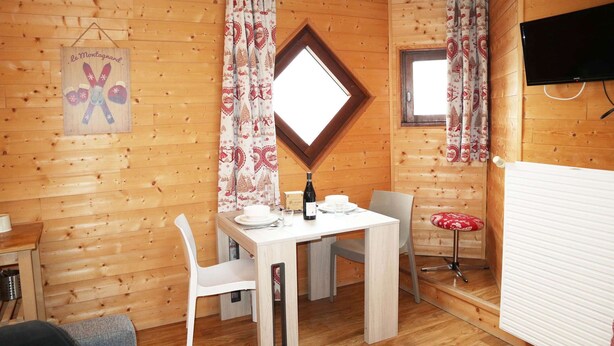 L’intérieur d’un chalet en bois comprend une table à manger, des chaises, un petit tabouret, un téléviseur et une fenêtre avec des rideaux.