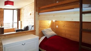 Un lit superposé avec une couverture rouge, une commode blanche à tiroirs et un mur en bois avec une fenêtre.