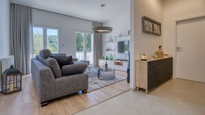A modern living room with a grey sofa, a coffee table, and a TV. There is a balcony with a view of greenery. - Happy Home Rab by Interhome (Rab/Rab)