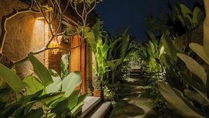 A well-lit pathway leading to a rustic building with a brick wall and wooden door, surrounded by lush tropical plants. - The Ulon Ubud Villa (Ubud)