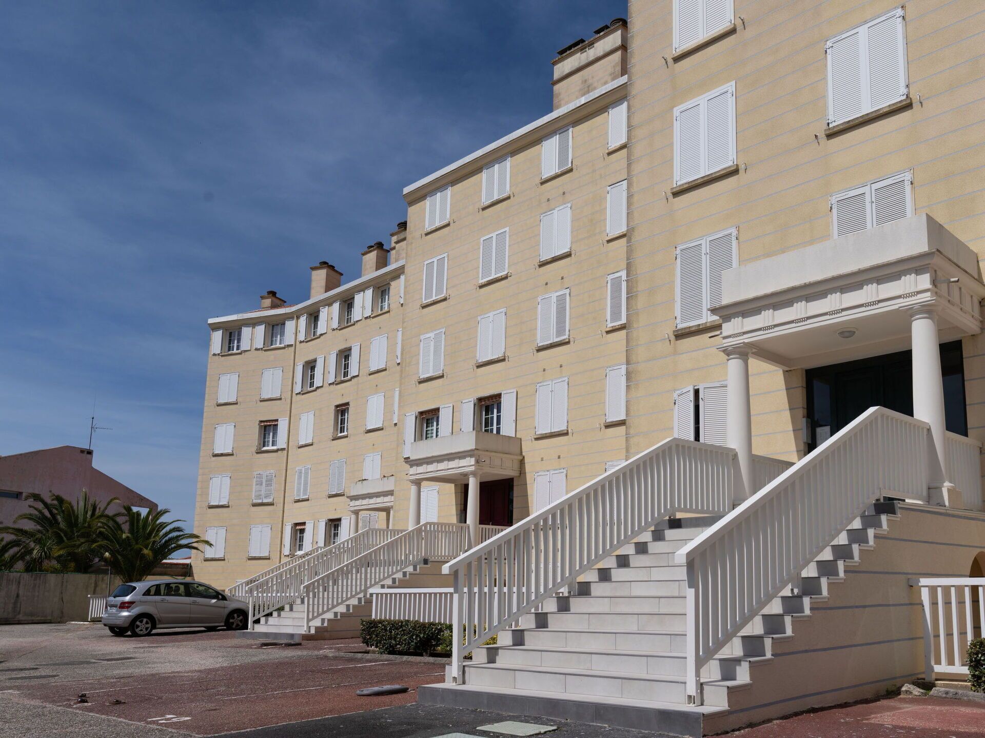 A multi-story building with a beige facade, white window shutters, and a central staircase with white railings.
