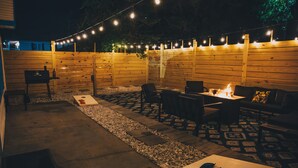 Un espace extérieur aménagé en patio, avec une clôture en bois, des guirlandes lumineuses, un foyer et du mobilier d’extérieur.