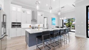 A modern kitchen with white cabinets, stainless steel appliances, and a large island with bar stools. - New Construction, Immaculate Canal Home; Wide Water Views, Pool, Spa, Dock & More ~Great Location! (Marco Island)