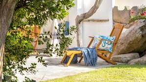 Une terrasse en bois avec une chaise et une couverture, entourée de verdure et bordée d’un mur blanc.