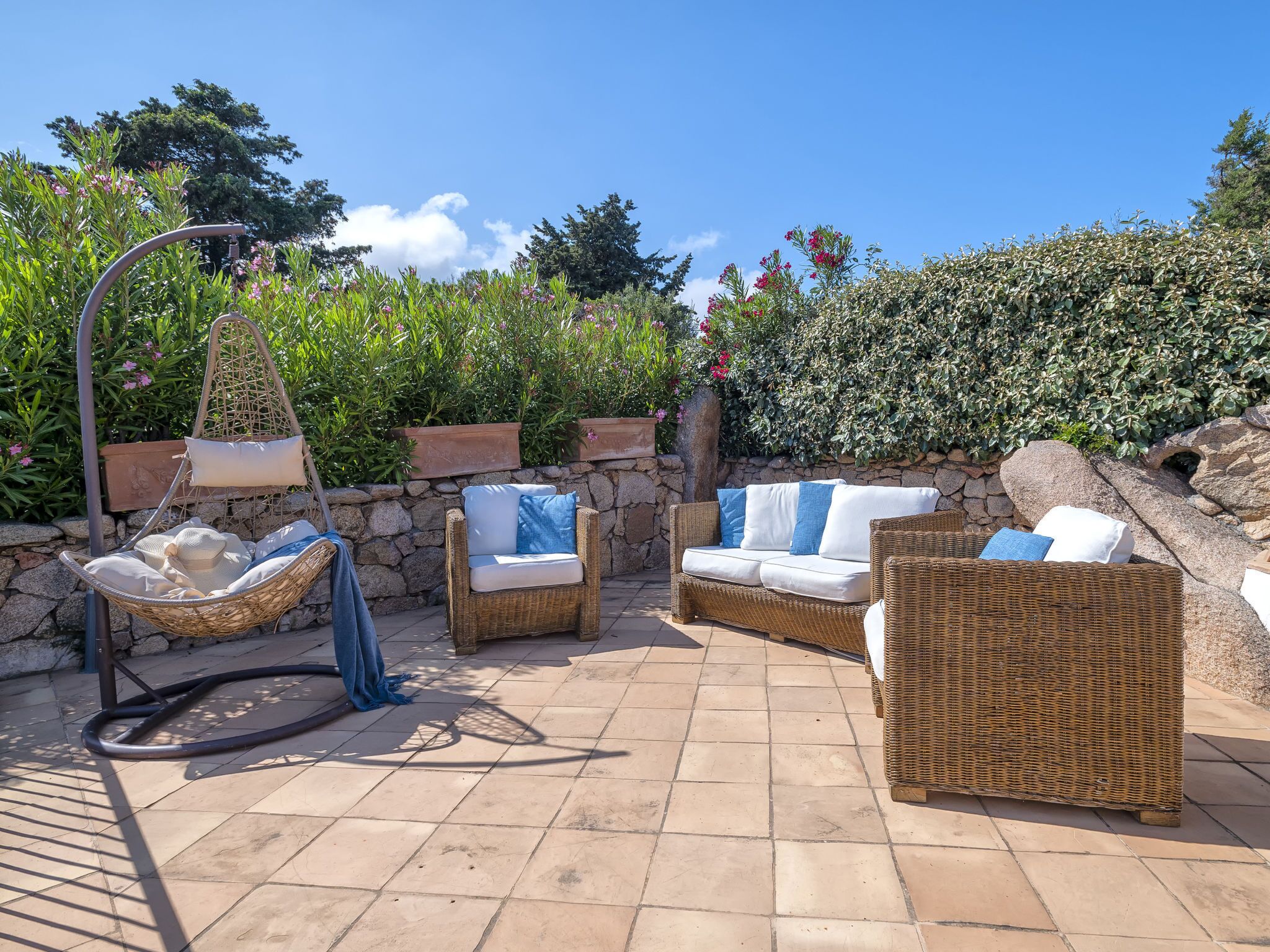 Un espace patio aménagé avec des meubles en osier, comprenant un fauteuil suspendu et deux canapés, le tout adossé à un mur en pierre et entouré d’une végétation luxuriante.