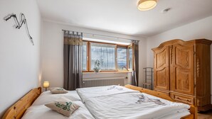 A bedroom with a double bed, a wooden wardrobe, a radiator, a window with curtains, and a wall-mounted light fixture. - Köberich Apartment with Mountain View, Private Terrace and Large Garden (Waging am See)