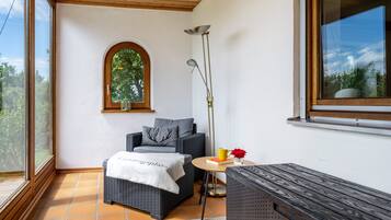 Una habitación con un ventanal grande, una mesita con un libro y una taza, un sofá con una manta y una lámpara de pie.