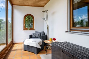 Une pièce avec une grande fenêtre, une petite table sur laquelle se trouvent un livre et une tasse, un canapé avec une couverture et une lampe sur pied.