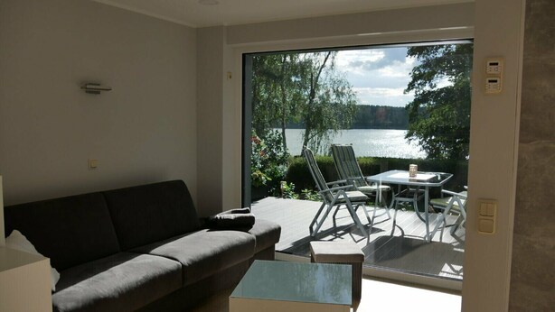 A modern living room with a sofa, a coffee table, and a view of a lake through a glass door. - Holiday Home 'Am Kiefernweg 17' with Private Terrace (Schwielochsee)