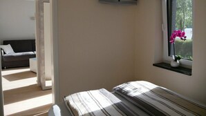 A bedroom with a bed, a TV mounted on the wall, a window with a flower vase, and a door leading to another room. - Holiday Home 'Am Kiefernweg 17' with Private Terrace (Schwielochsee)