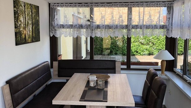 Una habitación con una mesa de madera, un banco con cojines negros, cortinas de encaje y un cuadro en la pared.