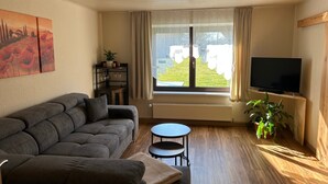 Una sala de estar con sofá, una mesita, televisión, ventana con cortinas y una planta.