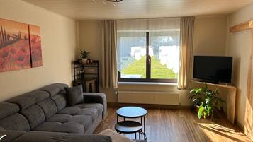 Ein Wohnzimmer mit einer Couch, einem kleinen Tisch, einem Fernseher, einem Fenster mit Vorhängen und einer Pflanze.