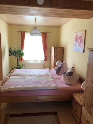 Un dormitorio con una cama, una mesita de noche, una lámpara, un radiador, una ventana con cortinas y un cuadro en la pared.