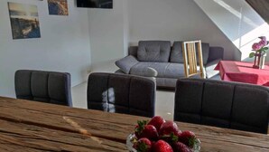 Een moderne woonkamer met een grijze bank, een houten eettafel met een schaal aardbeien en een rode tafelkleed.
