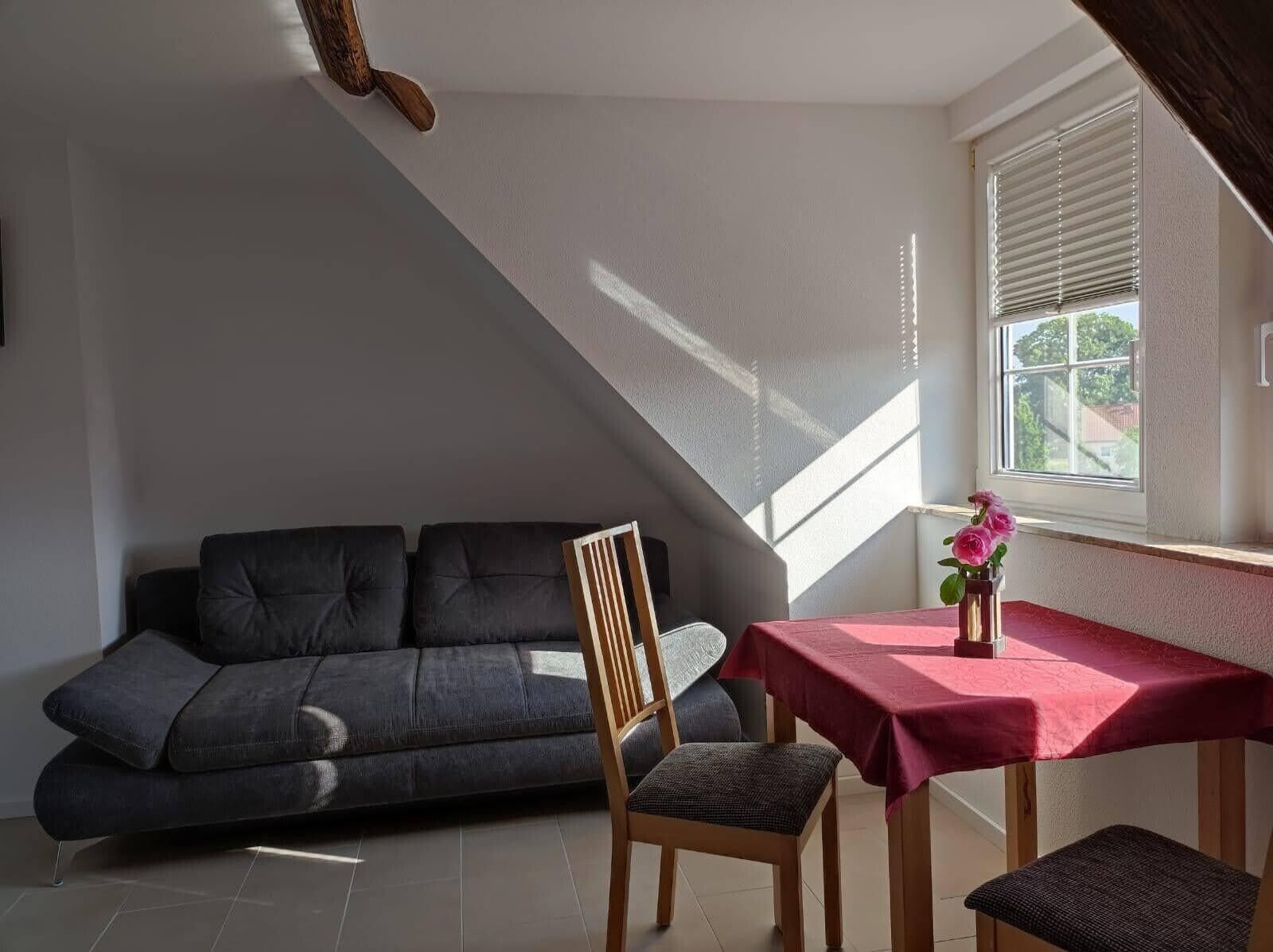 Une pièce moderne avec un canapé gris foncé, une table à manger en bois recouverte d’une nappe rouge, une chaise et une petite table sur laquelle est posé un vase de fleurs.