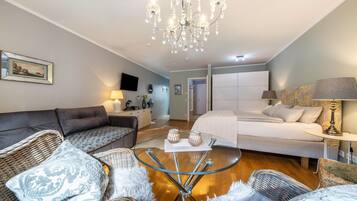 Quarto de hotel moderno com sofá, duas poltronas, mesa de centro de vidro, cama, lustre e televisão.