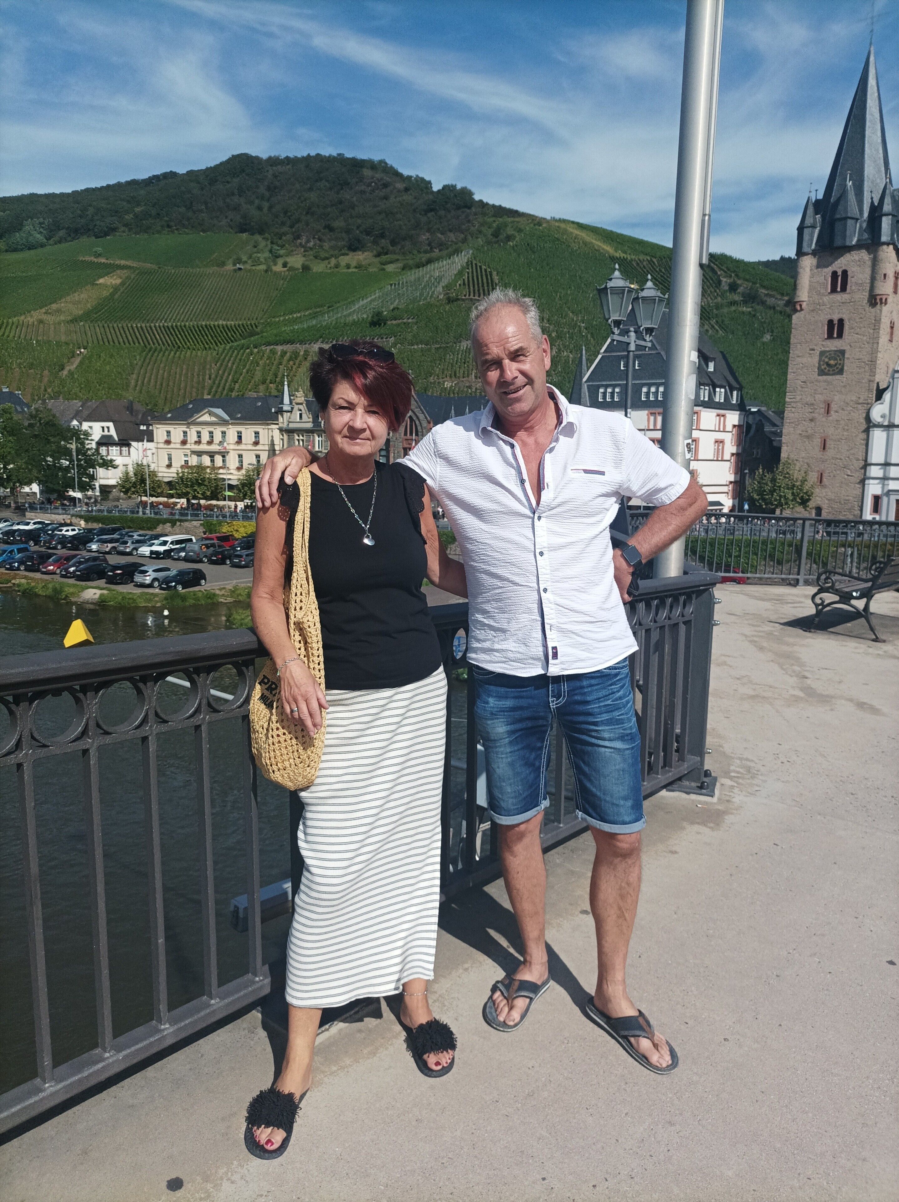 Um casal parado em uma ponte, com vista para um castelo e vinhedos ao fundo.