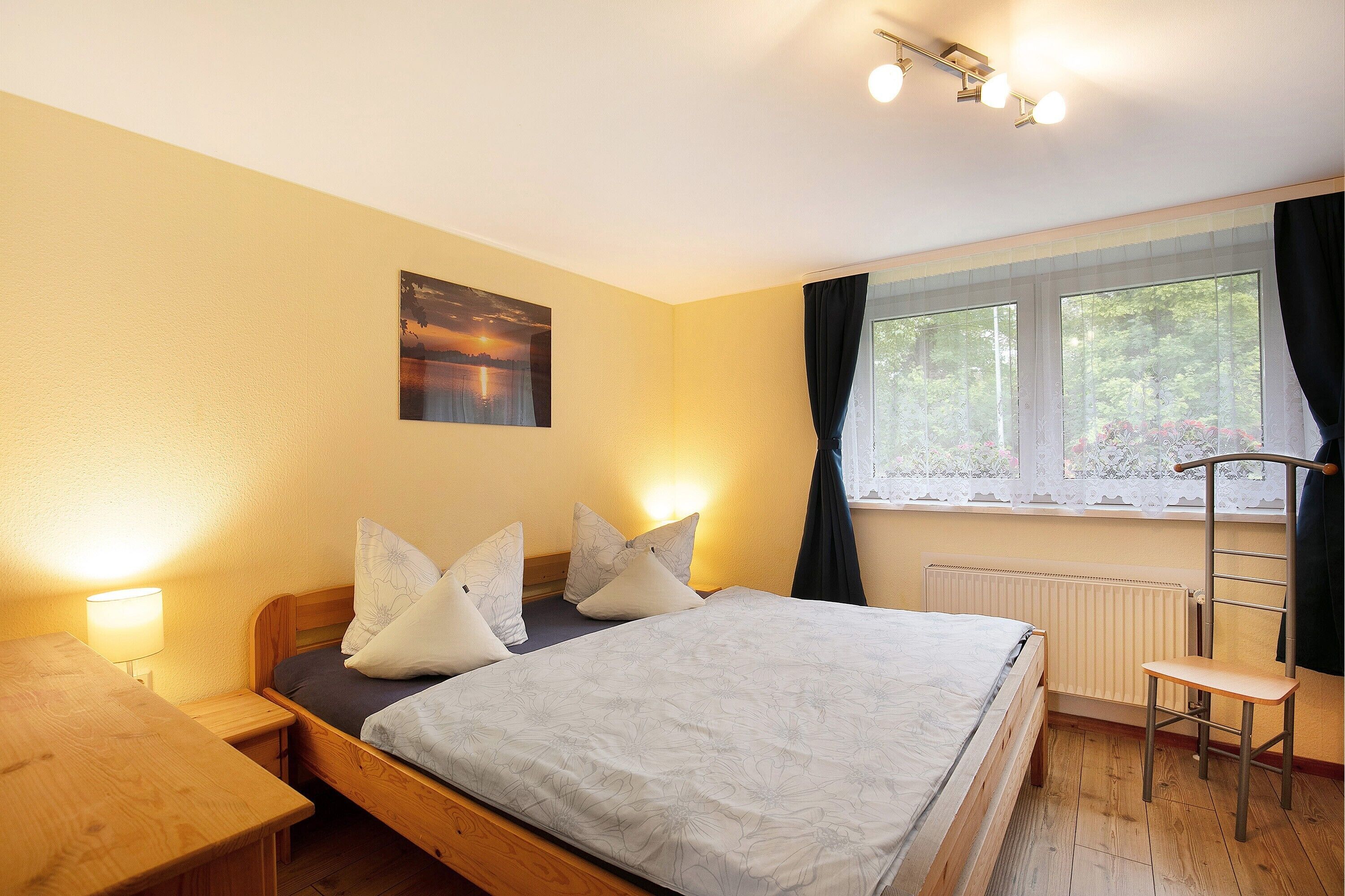Een slaapkamer met een bed, nachtkastje, lamp, raam met gordijnen, radiator en een stoel.