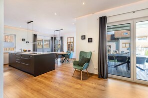 Eine moderne Küche mit einer zentralen Kochinsel, einem Essbereich mit Tisch und Stühlen sowie einer großen Glasschiebetür, die auf eine Terrassenfläche führt.