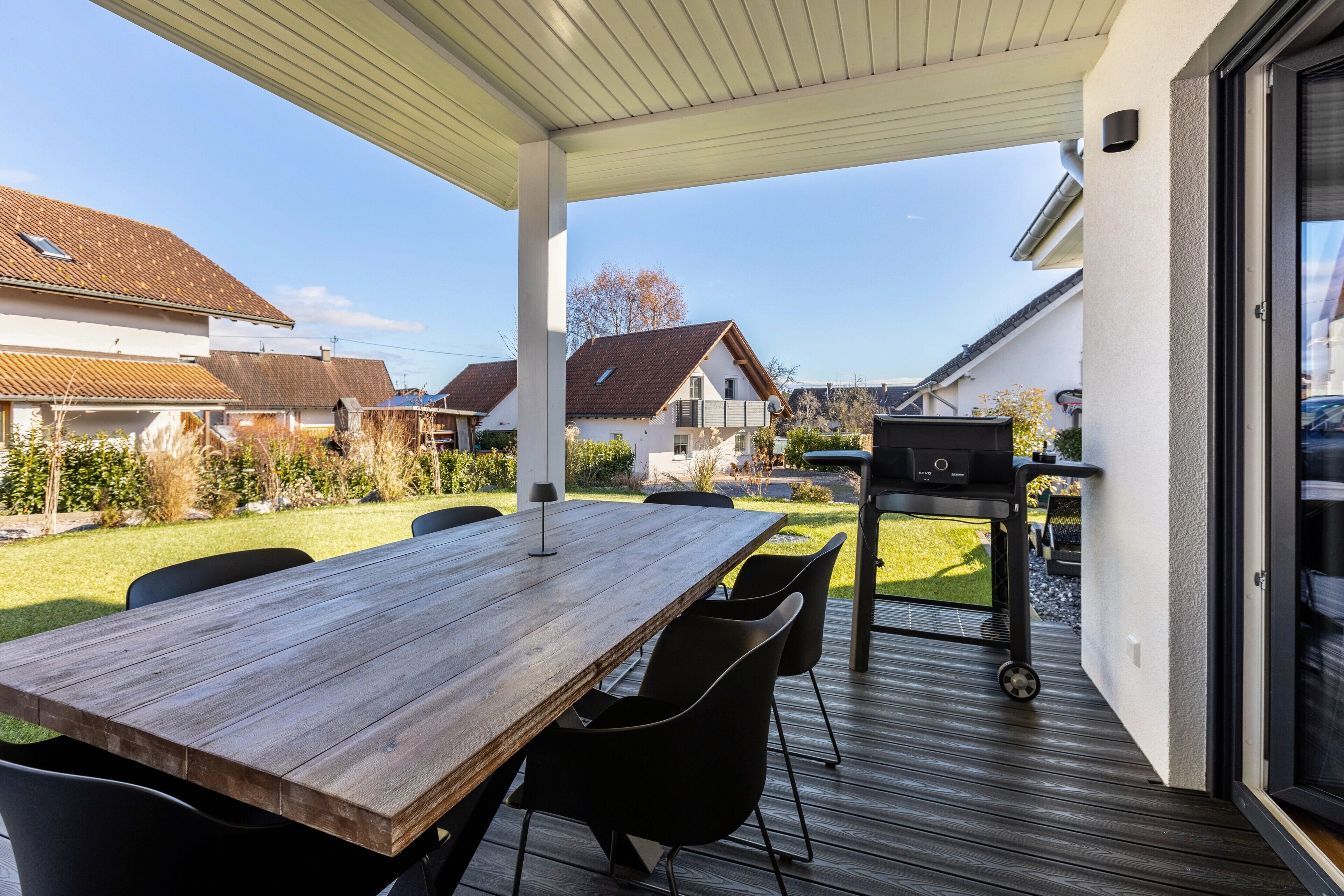 En inglasad uteplats med ett träbord och stolar, en grill och utsikt över de omkringliggande husen.