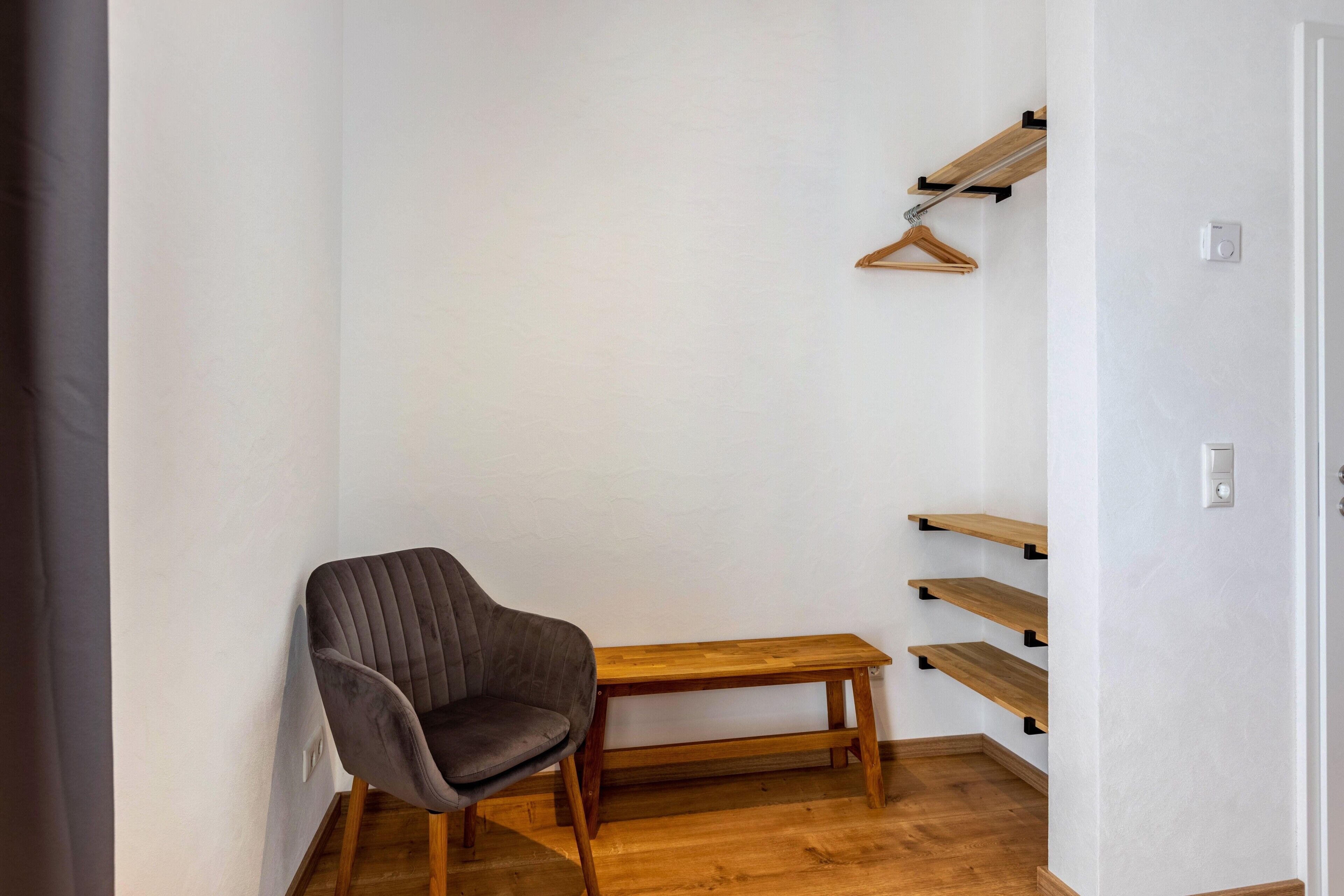 Une pièce d’angle avec un fauteuil à revêtement gris, un banc en bois et une étagère en bois fixée au mur, dotée de supports pour suspendre des vêtements.