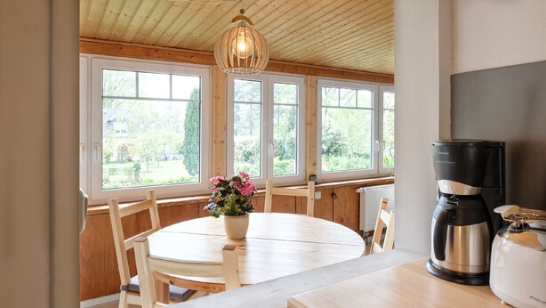 A kitchen with a round table, wooden chairs, a coffee maker, and a view of greenery through large windows. - Apartment 'Loschwitz' with Wi-Fi (Dresden)
