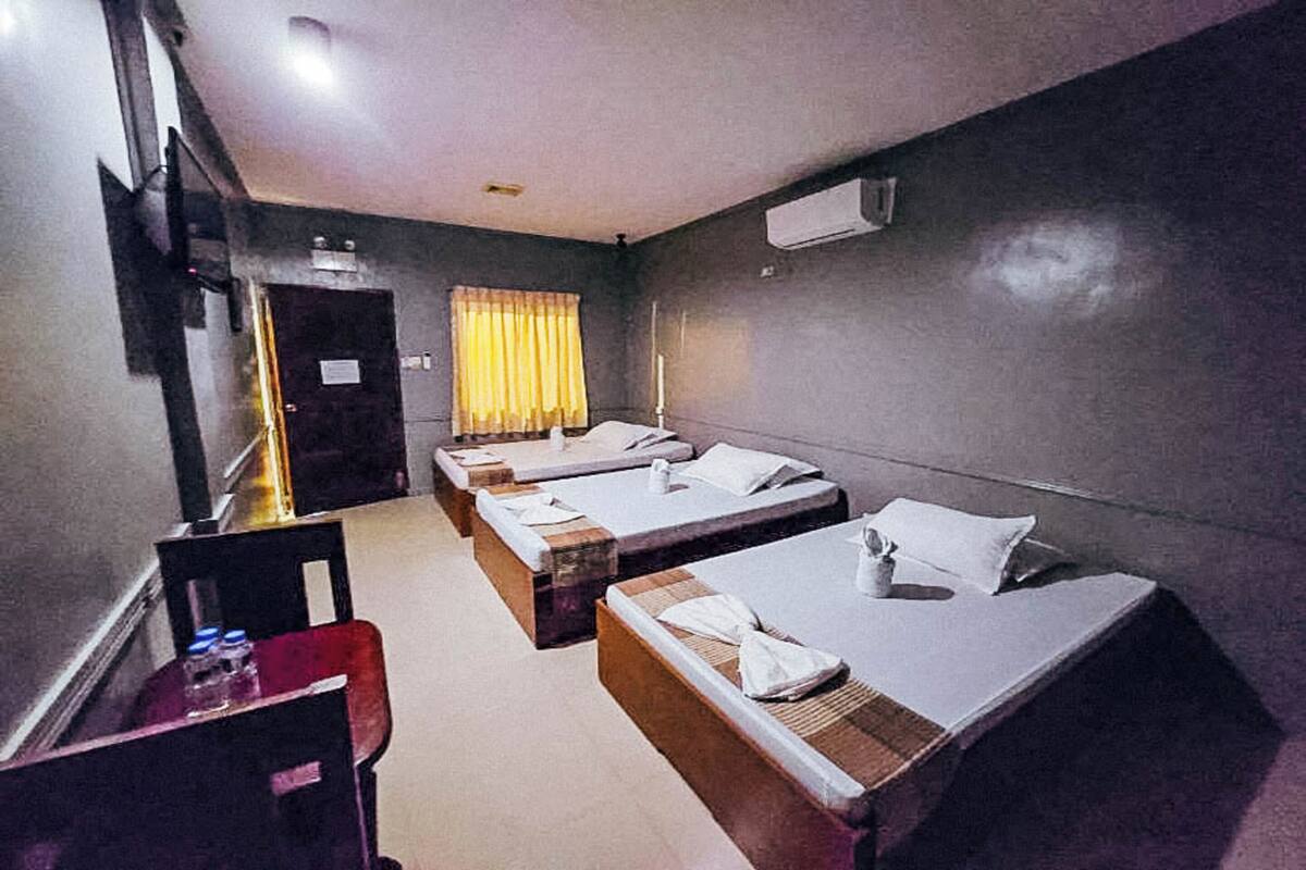 Ein Hotelzimmer mit drei Einzelbetten, einem Schreibtisch mit Stuhl, einem Fernseher und einer Klimaanlage.