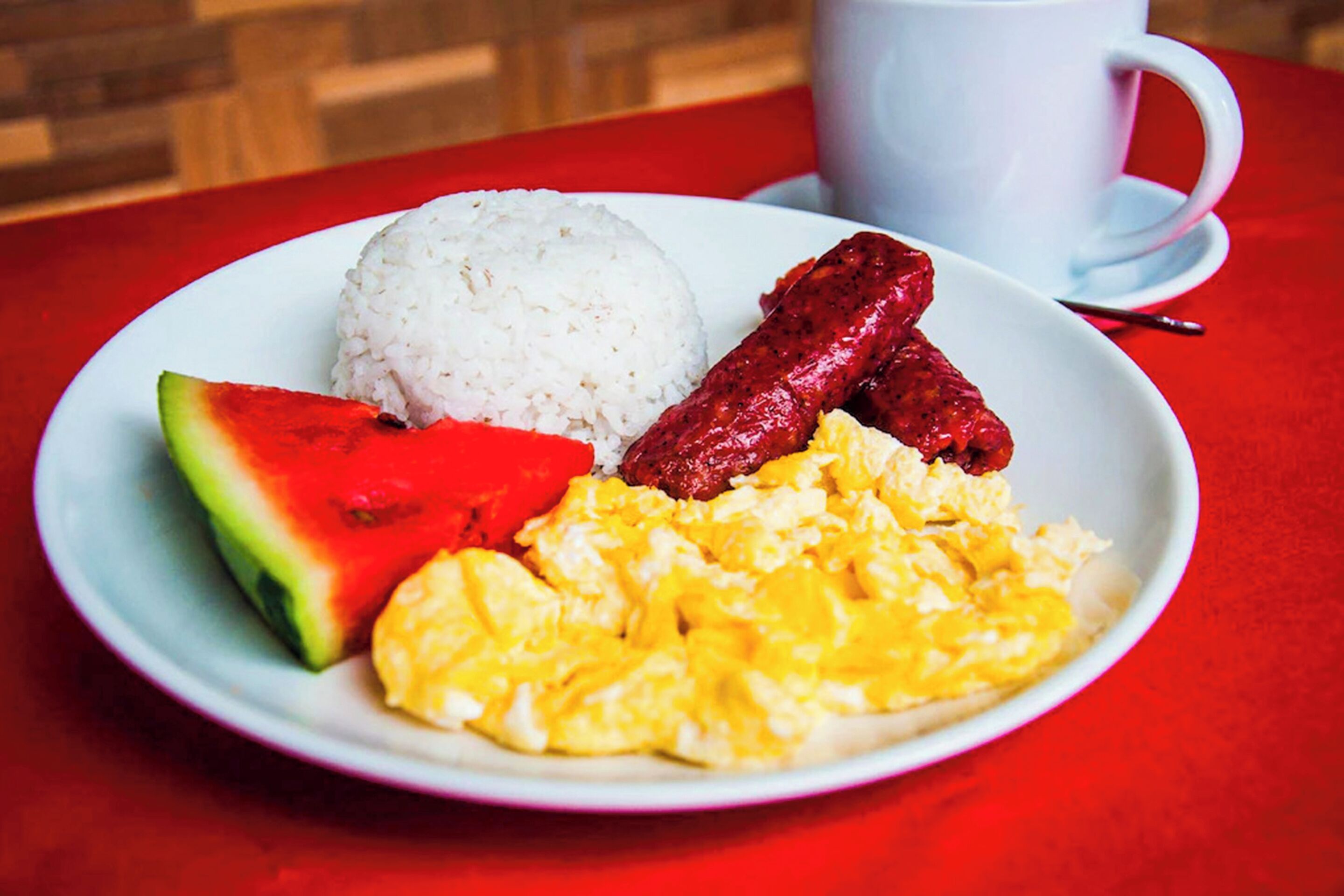 Un plato con arroz, salchicha, huevos revueltos y una porción de sandía sobre una mesa roja.