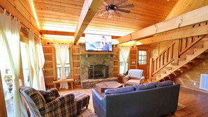 A log cabin interior with a stone fireplace, a television, plaid and blue sofas, and a wooden staircase.