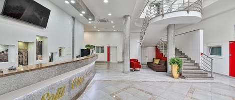 A spacious lobby with a reception desk, a staircase, and a large TV screen.