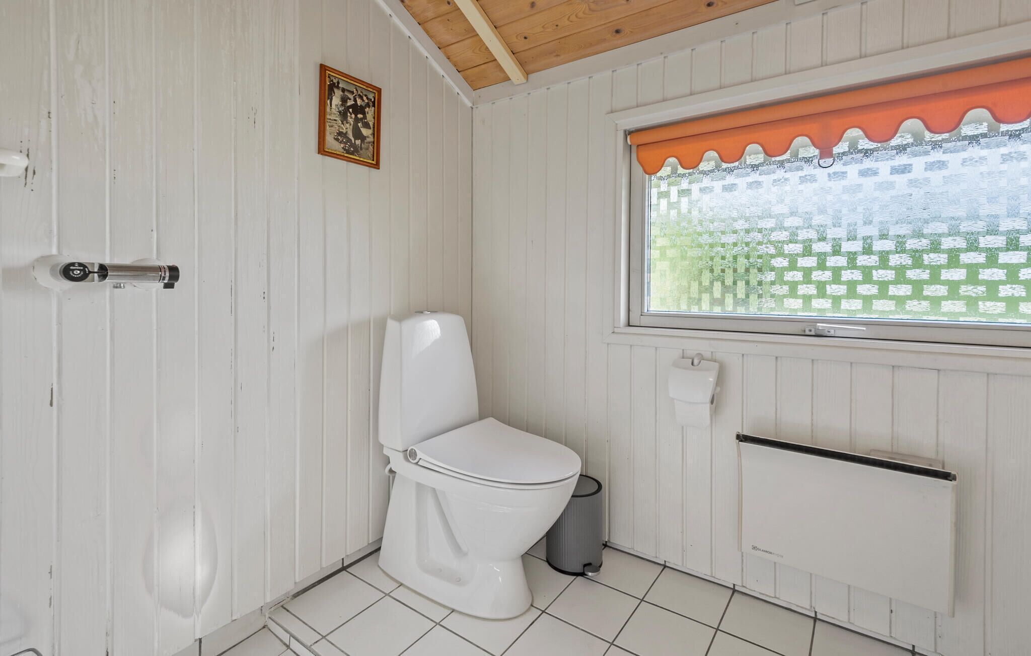 Un baño pequeño con paneles blancos, un inodoro, una ventana con una persiana verde a cuadros y un pequeño cuadro en la pared.