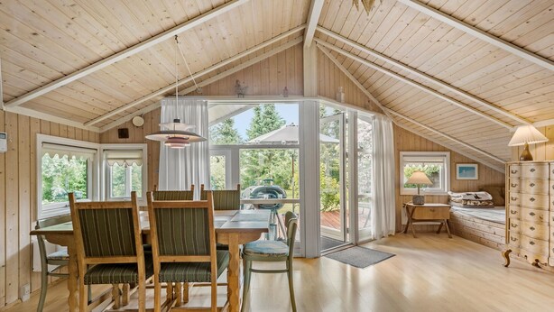 Ein gemütliches Dachzimmer mit Holzbalkendecke, einem Esstisch mit Stühlen, einem Fenster mit Vorhängen und einer Lampe.