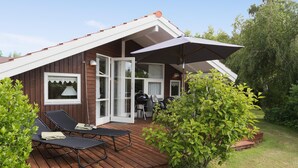 Eine Holzhütte mit einer Terrasse, zwei Liegestühlen, einem Grill und einem großen Sonnenschirm.