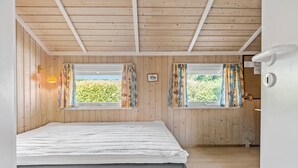 Ein Schlafzimmer mit Holzboden, zwei mit Vorhängen versehenen Fenstern, einem Bett und einem kleinen Nachttisch.