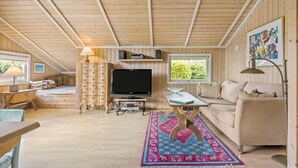 Ein gemütliches Blockhauss-Interieur mit Holzbalkendecke, einem Fernseher, einer Couch, einem Esstisch und einem gemusterten Teppich.