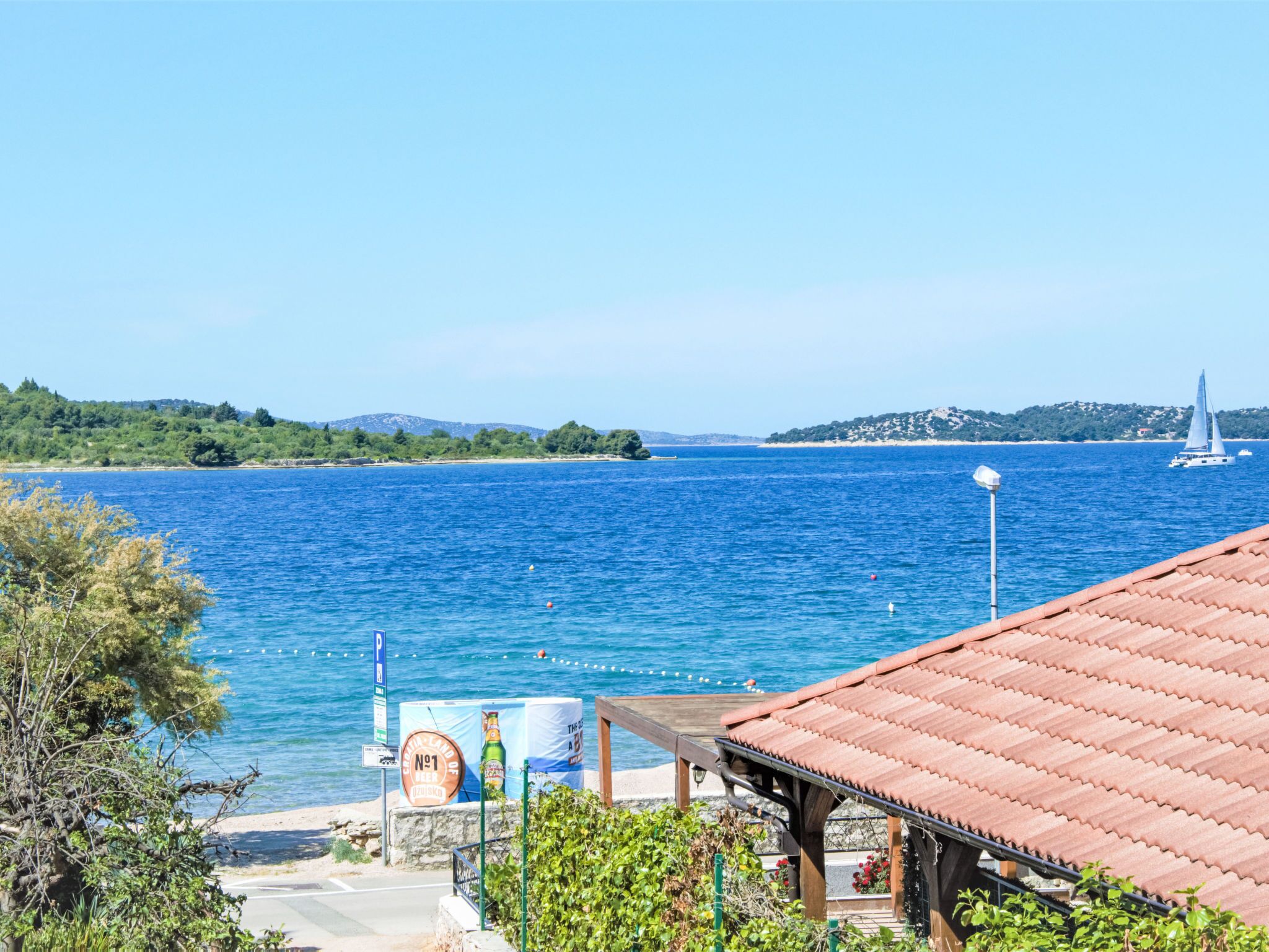 A coastal view with a clear blue sea, a sailboat, and a building with a tiled roof.