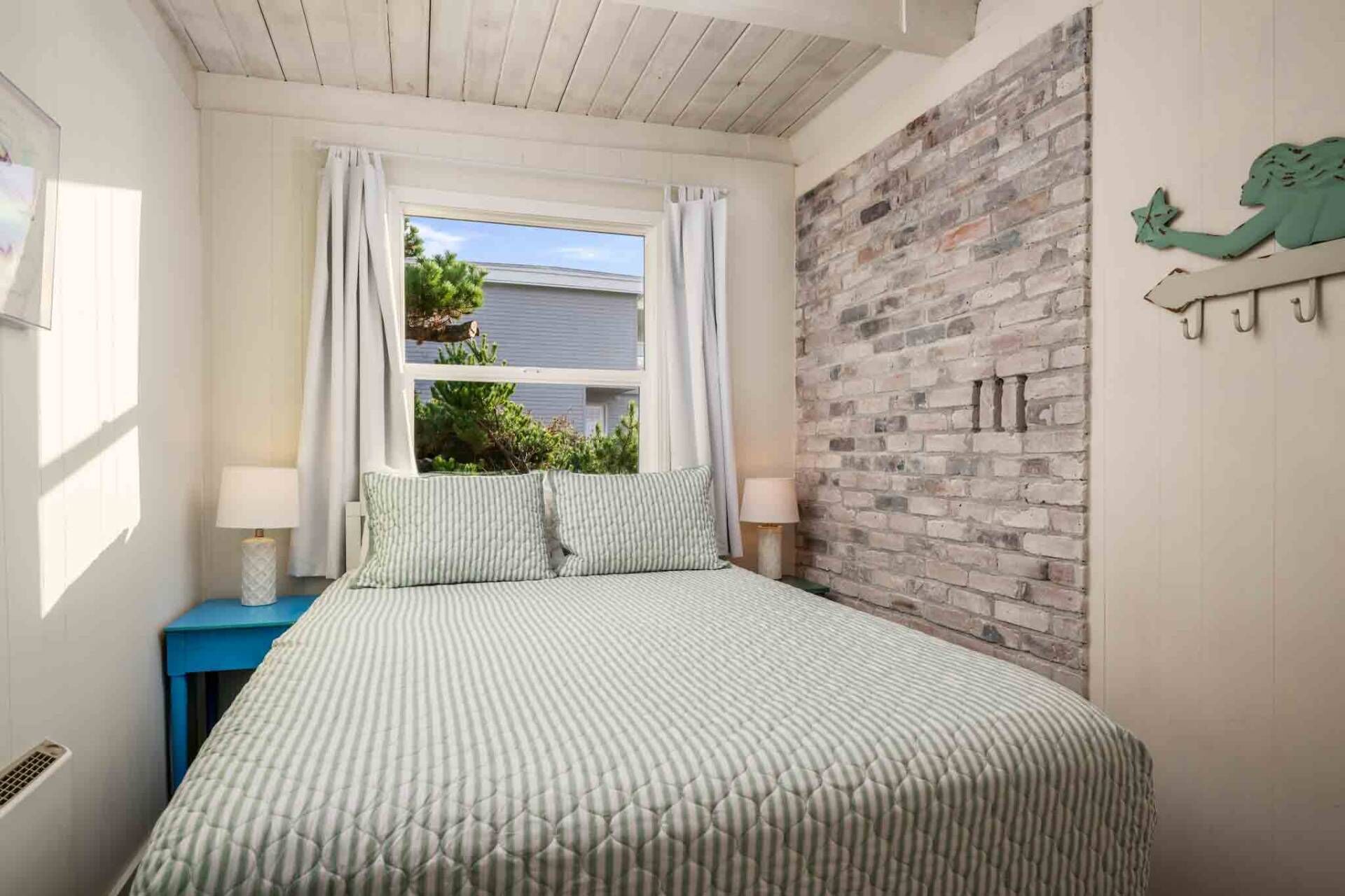 Een slaapkamer met een bed, een raam met gordijnen, een bakstenen muur en een kleine tafel met een lamp.